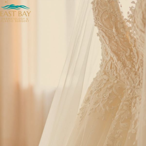 Close-up of an ivory lace wedding dress with intricate embroidery and sheer veil, with the East Bay Dermatology & Plastic Surgery logo in the corner.