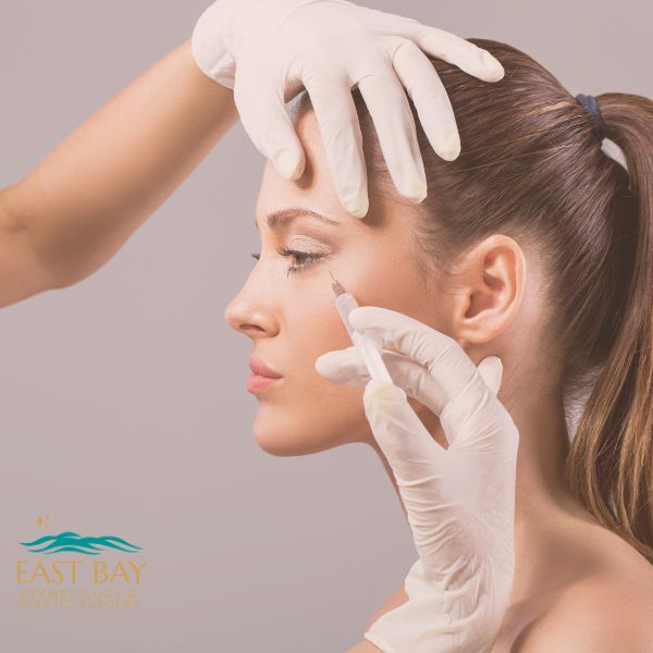 A woman receives an injection near her eye from a gloved professional. The logo "East Bay" is visible in the lower left corner.