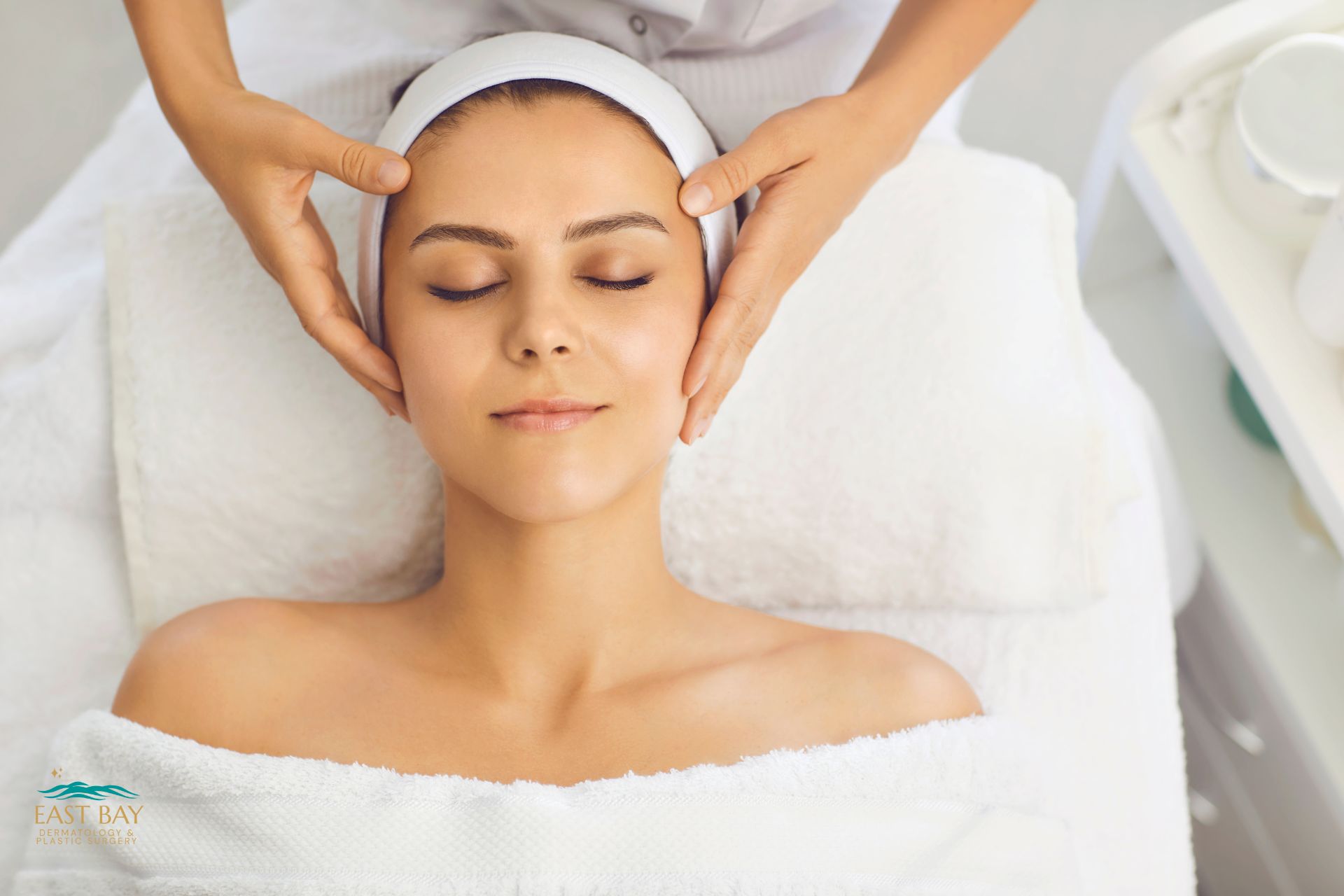 A person with a headband and towel receives a facial massage while lying on a treatment table in a spa setting.