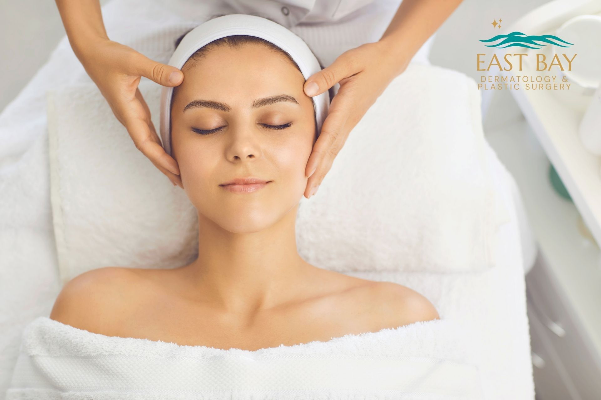 A woman lies on a spa bed with her eyes closed, receiving a facial massage. She is covered with a towel and wears a white headband. The East Bay Dermatology & Plastic Surgery logo highlights expert skin care in Bellmore.