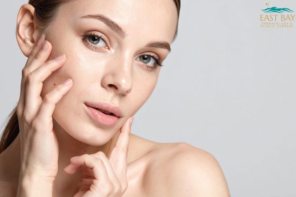 A woman with clear skin touches her face gently with both hands against a light background. The East Bay Dermatology & Plastic Surgery logo is visible in the top right corner.