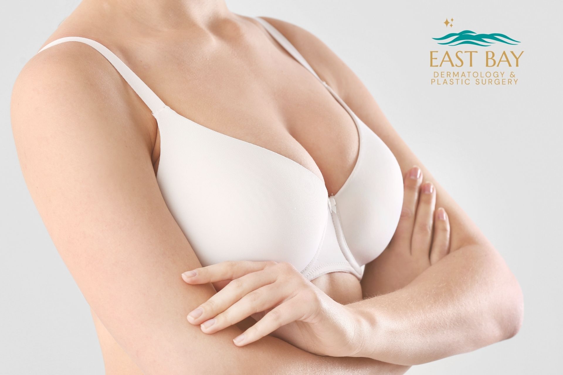 A woman in a white bra crosses her arms over her torso, reflecting confidence after breast augmentation in Bellmore NY. The logo for East Bay Dermatology & Plastic Surgery is visible in the top right corner.
