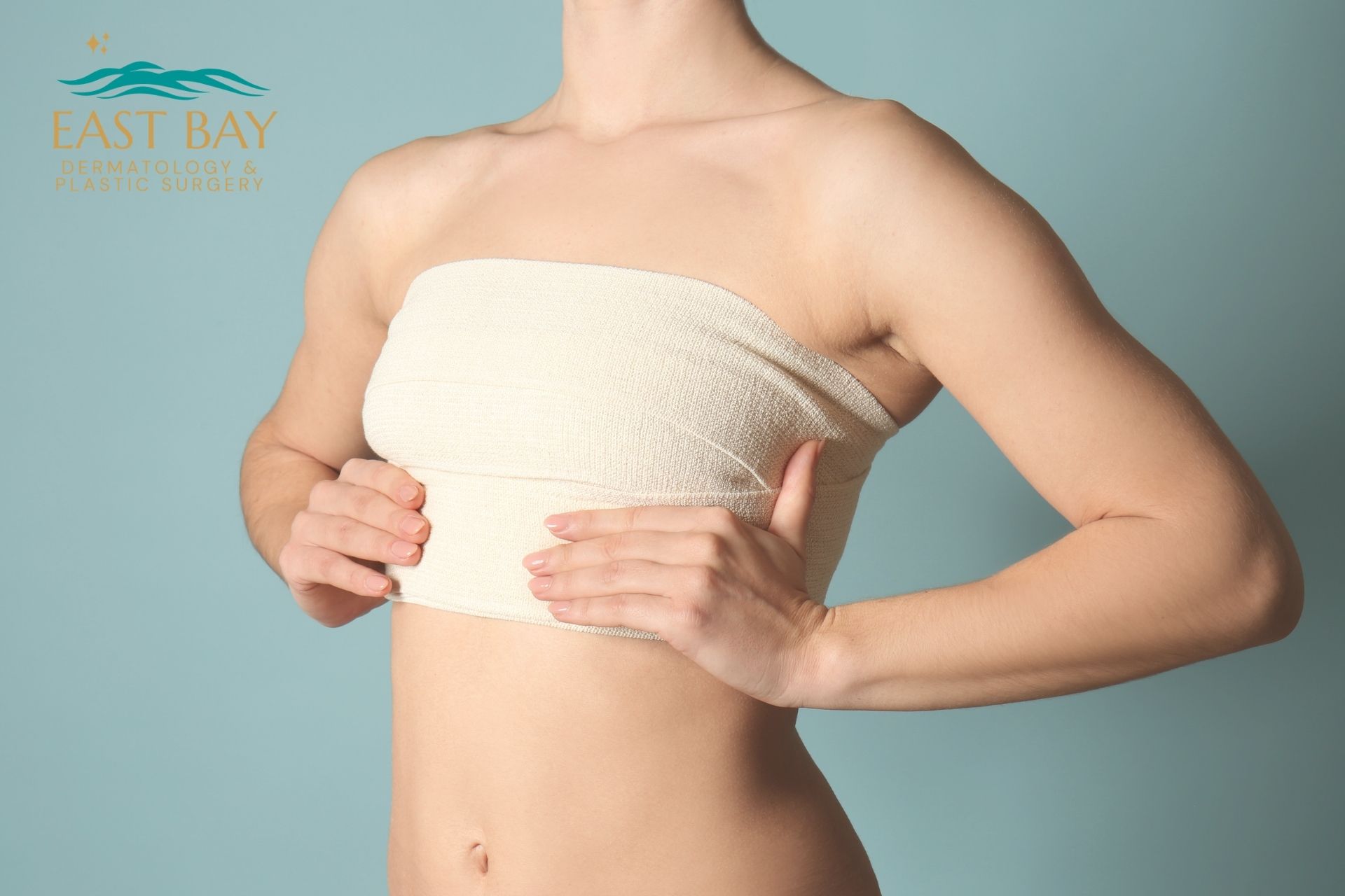 A woman with a bandage around her chest places her hands below her breasts, highlighting the results of breast augmentation in Bellmore NY; East Bay Dermatology & Plastic Surgery logo is in the corner.