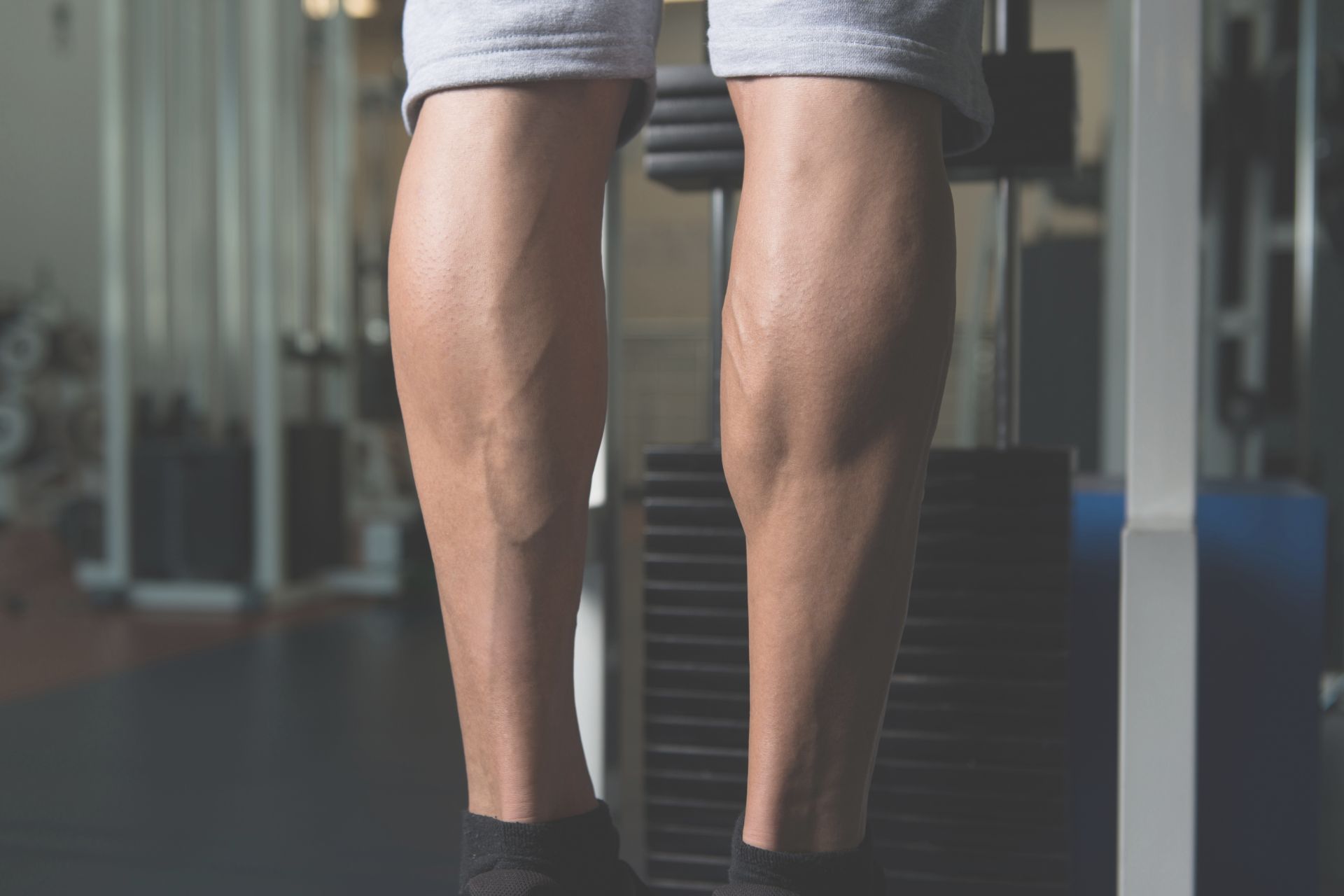 Close-up of a person's lower legs and calves, standing in a gym, wearing shorts and socks, with gym equipment visible in the background—ideal for illustrating fitness before or after surgical treatments.