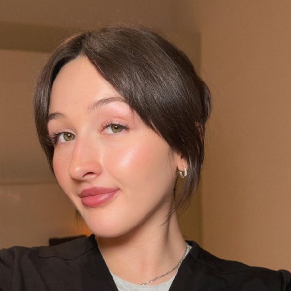 A young woman with fair skin and straight brown hair smiles slightly, wearing a black top and small hoop earrings, posing indoors against a beige background.
