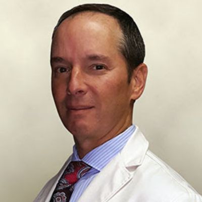 A man with short dark hair wearing a white lab coat, light blue shirt, and patterned tie, posing against a plain light background.