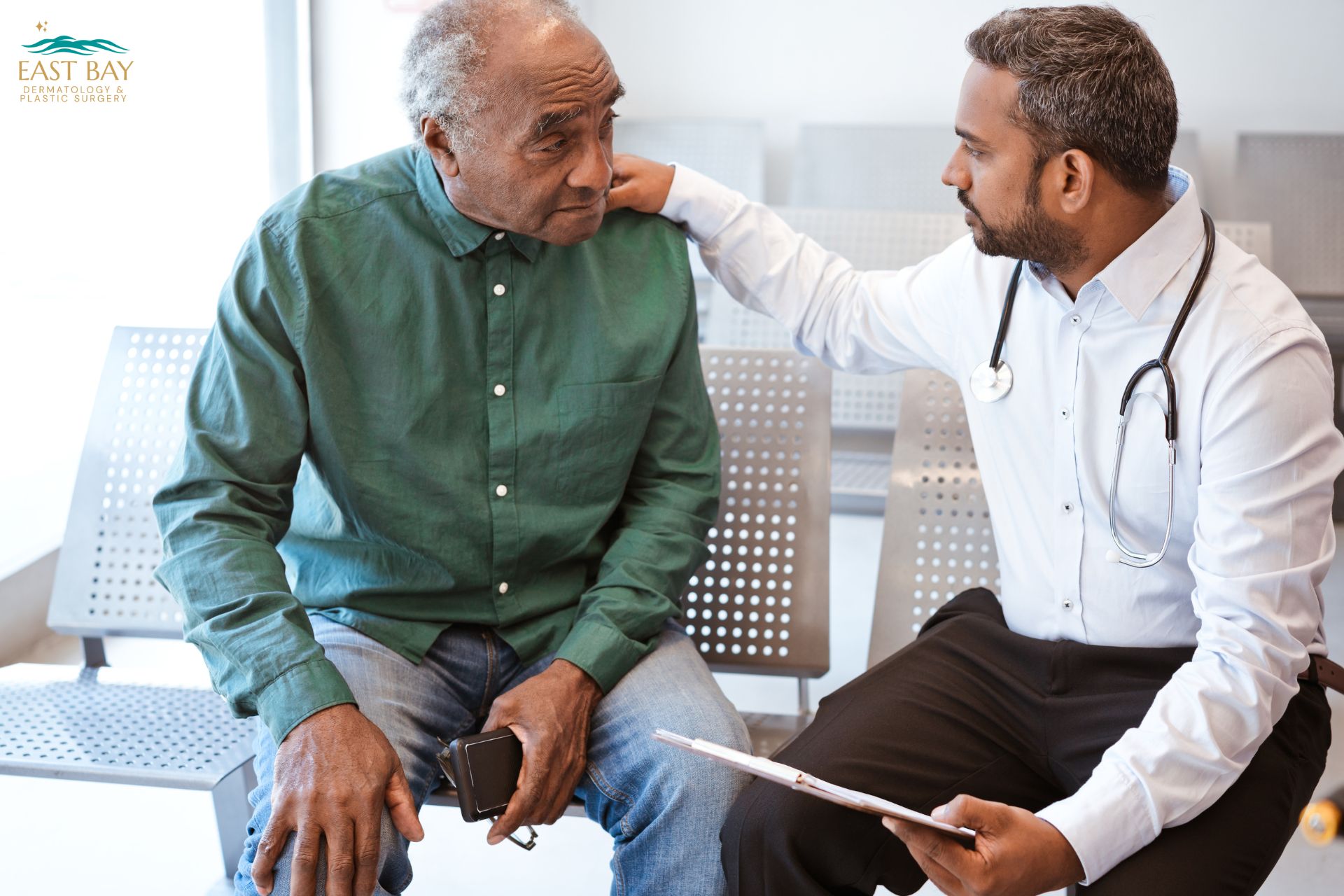 A doctor with a stethoscope on his neck comforts an elderly man in a waiting room, touching his shoulder and holding a clipboard.