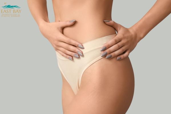 A woman in beige underwear poses with her hands on her hips against a plain background. The logo "East Bay Aesthetic Surgery" appears in the top left corner.