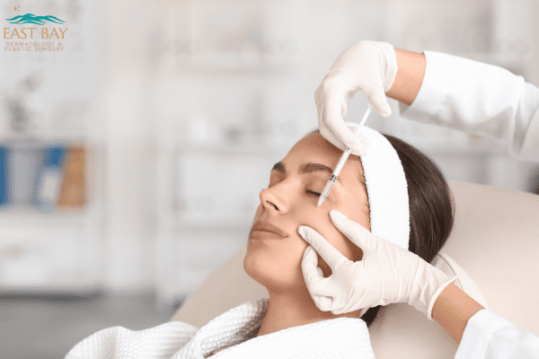 A person receives a cosmetic injection in the forehead area while lying on a medical chair; a healthcare professional wears gloves. East Bay Dermatology & Plastic Surgery logo in the corner.