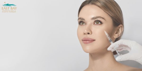 A woman with clear skin receives a cosmetic injection in her cheek from a gloved hand; East Bay Dermatology logo is visible in the corner.