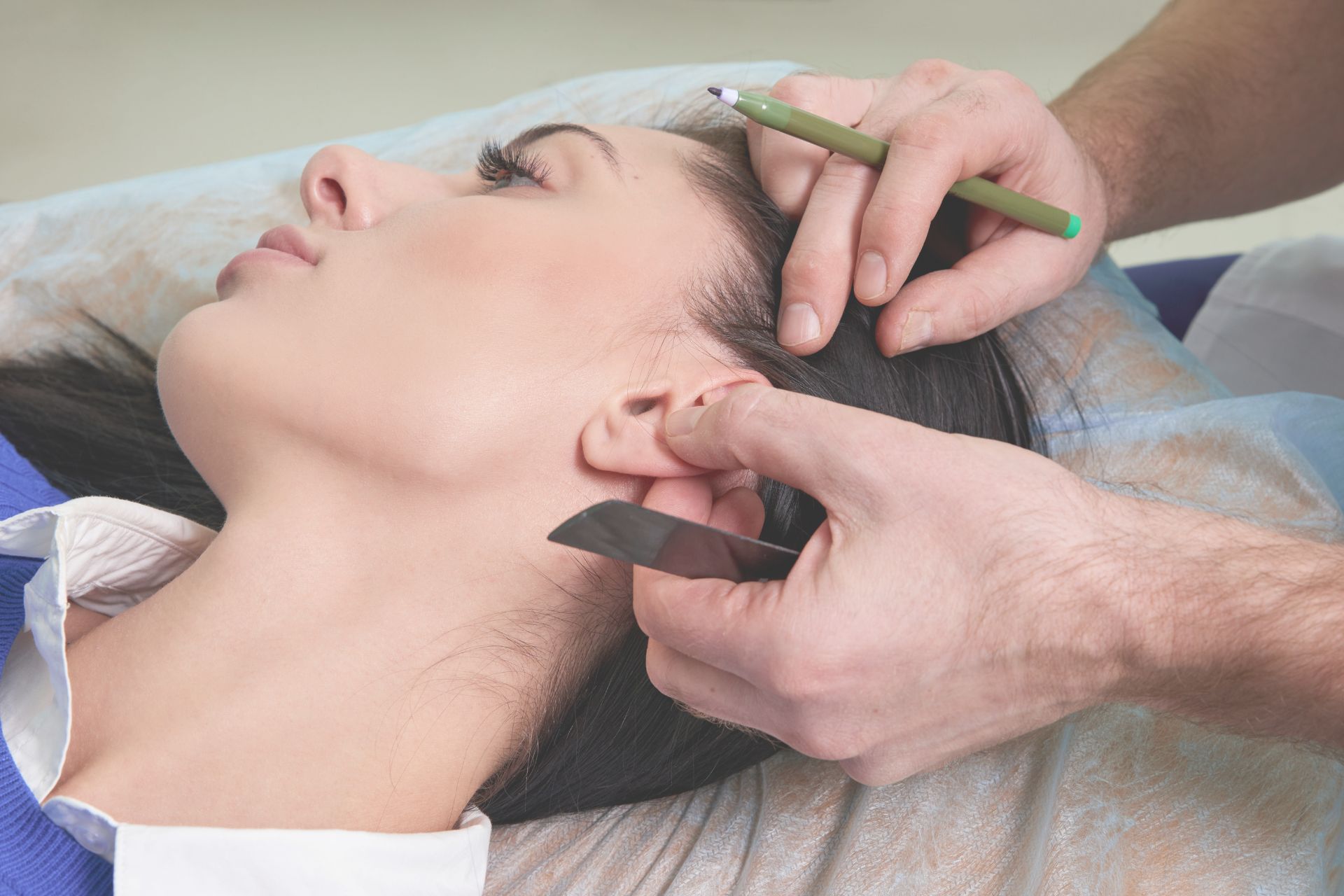 Person lying on their side while a medical professional marks their ear with a green marker and holds a ruler, likely preparing for a cosmetic or surgical procedure.