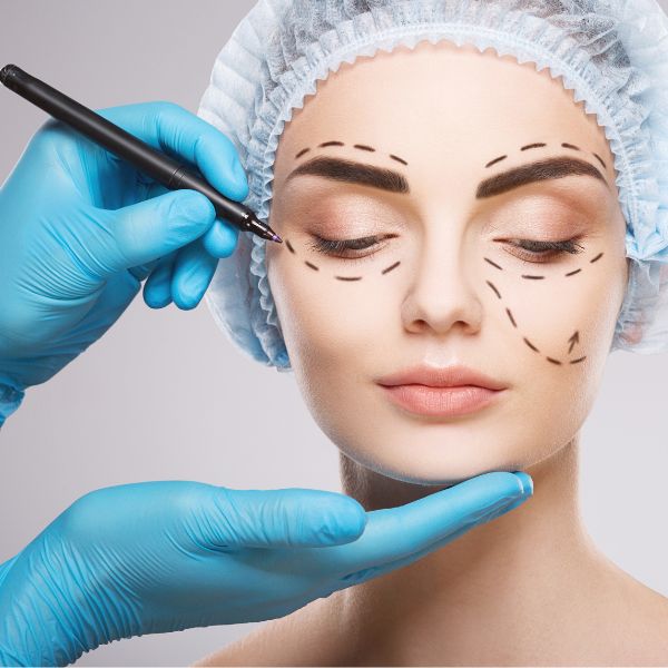 A person wearing a surgical cap has dotted lines drawn on their face as a gloved hand holds a marker near their eye, indicating cosmetic surgery planning.
