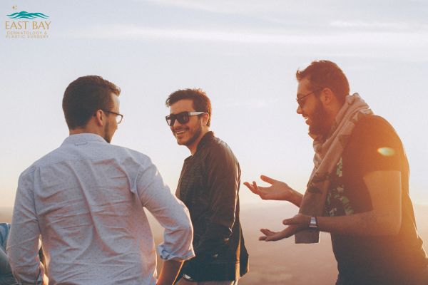 Three men stand outdoors in sunlight, smiling and talking together. One man gestures with his hands, and all appear relaxed. A logo is visible in the top left corner.