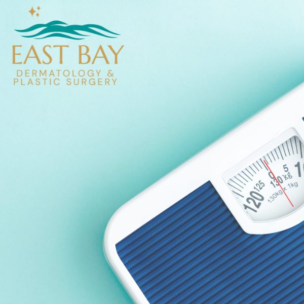 A corner of a blue analog bathroom scale displaying numbers, with the East Bay Dermatology & Plastic Surgery logo in the top left corner on a light blue background.